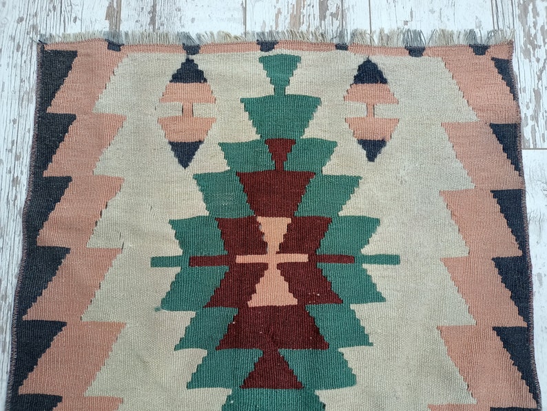 K&ouml;nnte beinhalten: Ein gewebter Teppich mit einem geometrischen Muster in Beige-, Rosa-, Gr&uuml;n- und Dunkelblaut&ouml;nen. Das Design zeigt sich wiederholende Dreiecks- und Diamantformen, die eine strukturierte Oberfl&auml;che erzeugen. Der Teppich hat einen Fransenrand.