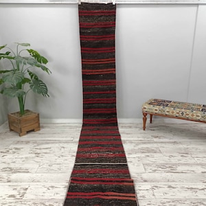 May include: A long, narrow red and black striped rug with a distressed white wood floor background.