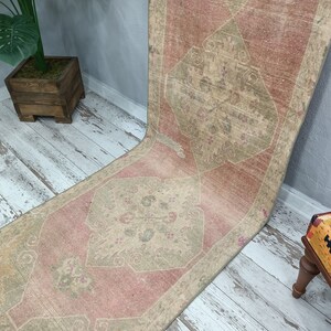 May include: A vintage runner rug with a faded pink and beige colour scheme. The rug has a traditional geometric pattern with a central diamond shape.