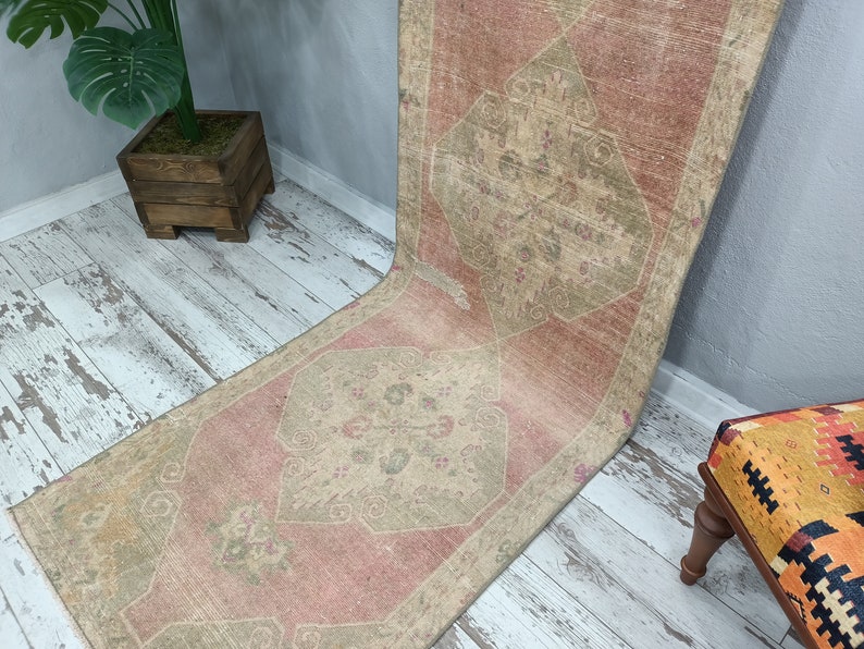 May include: A vintage runner rug with a faded pink and beige colour scheme. The rug has a traditional geometric pattern with a central diamond shape.