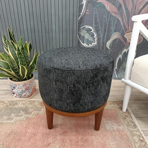 Può includere: Un pouf in tessuto nero e grigio con motivi e gambe in legno. Il pouf è su un tappeto rosa e beige.