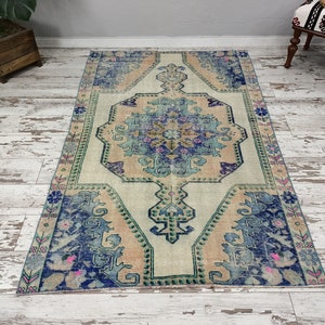 K&ouml;nnte beinhalten: Ein Vintage-Teppich mit einem verblassten Blumenmuster in Blaut&ouml;nen, Rosa und Gr&uuml;n. Der Teppich hat einen cremefarbenen Hintergrund und ein zentrales Medaillon mit einem geometrischen Muster.
