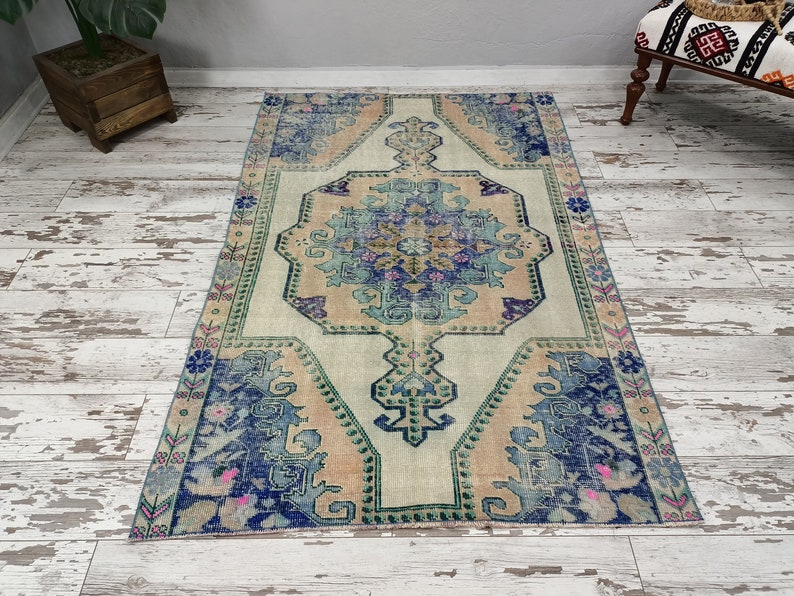 K&ouml;nnte beinhalten: Ein Vintage-Teppich mit einem verblassten Blumenmuster in Blaut&ouml;nen, Rosa und Gr&uuml;n. Der Teppich hat einen cremefarbenen Hintergrund und ein zentrales Medaillon mit einem geometrischen Muster.
