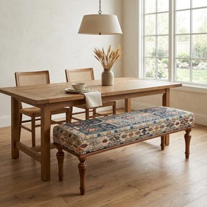 a cozy dining area with a wooden table, two chairs, and a bench with a colorful patterned seat. The table is set with a white bowl and a vase of dried flowers, creating a warm and inviting atmosphere. The room has a large window that provides a view of the outdoors, and the wooden floor adds a touch of rustic charm to the space.