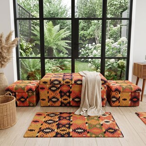 May include: A living room scene featuring a patterned rug, storage ottomans, and a blanket. The rug and ottomans have a geometric design in shades of orange, black, and green. A large window with a view of greenery is in the background.