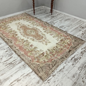 Op de afbeelding: Een vintage tapijt met een bloemenpatroon in tinten bruin, roze en cr&egrave;me. Het tapijt heeft een verweerde uitstraling en ligt op een witgeschilderde houten vloer.