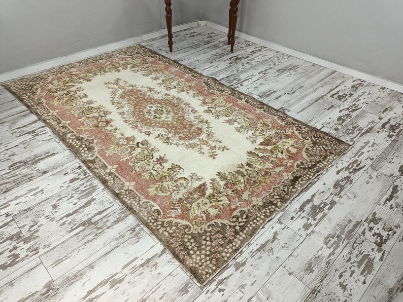 Op de afbeelding: Een vintage tapijt met een bloemenpatroon in tinten bruin, roze en cr&egrave;me. Het tapijt heeft een verweerde uitstraling en ligt op een witgeschilderde houten vloer.