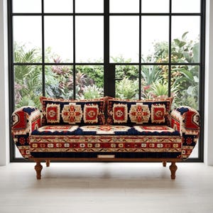 Könnte beinhalten: Ein gemustertes Sofa mit Holzrahmen und vier Beinen. Das Sofa hat ein rot-, blau- und cremefarbenes Design mit passenden Wurfkissen. Das Sofa steht vor einem großen Fenster mit Blick auf einen üppigen Garten.