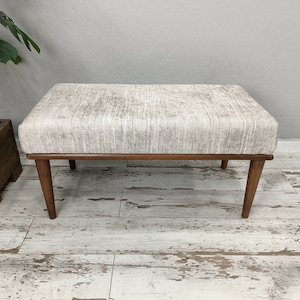 Può includere: Una panca imbottita di colore grigio chiaro con una struttura in legno e quattro gambe. La panca è appoggiata su un pavimento in legno chiaro.