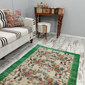 May include: A white and black striped sofa with a colorful patterned pillow sits in front of a green and cream floral rug. A small wooden table with a patterned top and a drawer sits next to a small cream and brown patterned ottoman.