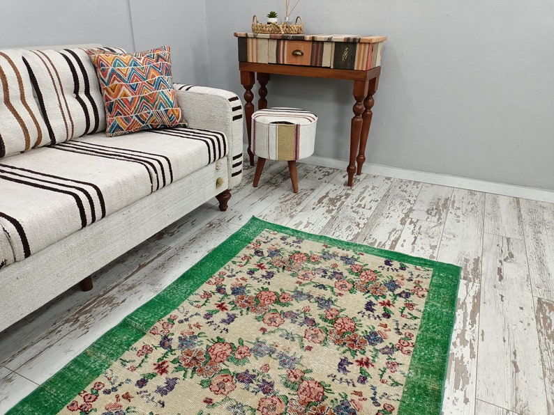 May include: A white and black striped sofa with a colorful patterned pillow sits in front of a green and cream floral rug. A small wooden table with a patterned top and a drawer sits next to a small cream and brown patterned ottoman.