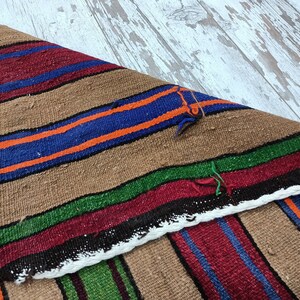 May include: A brown, woven rug with colorful stripes in shades of red, blue, green, and orange. The rug is folded in half, showing the pattern on both sides.