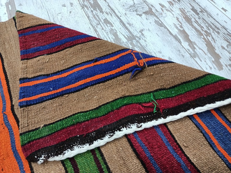 May include: A brown, woven rug with colorful stripes in shades of red, blue, green, and orange. The rug is folded in half, showing the pattern on both sides.