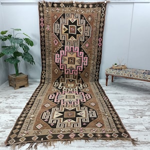May include: A long, rectangular rug with a brown, beige, and pink geometric pattern. The rug has a fringe on the bottom edge.