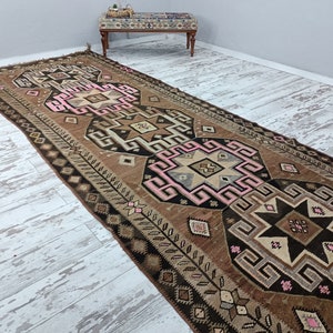 May include: A brown and pink patterned runner rug with geometric designs. The rug has a fringe on the ends and is made of wool.