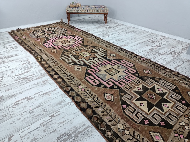 May include: A brown and pink patterned runner rug with geometric designs. The rug has a fringe on the ends and is made of wool.