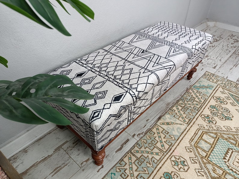 May include: A white and black patterned ottoman with wooden legs. The ottoman is sitting on a white wood floor with a patterned rug in the background.