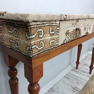 Op de afbeelding: Een houten consoletafel met een blad van patchwork stof. De stof is een mix van beige, bruin en wit met een geometrisch patroon. De tafel heeft gedraaide poten en een enkele lade met een messing handvat.