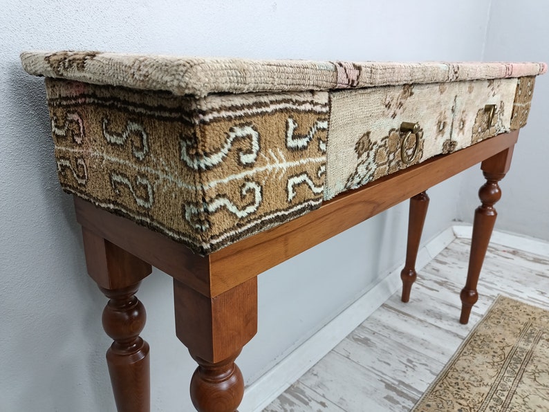 Op de afbeelding: Een houten consoletafel met een blad van patchwork stof. De stof is een mix van beige, bruin en wit met een geometrisch patroon. De tafel heeft gedraaide poten en een enkele lade met een messing handvat.