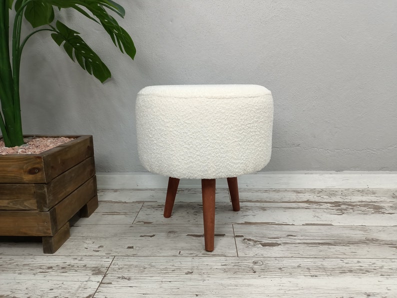 Puede incluir: Otomana blanca y esponjosa con cuatro patas de madera. Las patas son de color marr&oacute;n rojizo. La otomana est&aacute; sobre un suelo de madera clara.