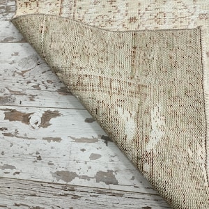 Puede incluir: Primer plano del reverso de una alfombra tejida beige y marr&oacute;n. La alfombra tiene un aspecto desgastado y est&aacute; doblada sobre un suelo de madera blanqueada.