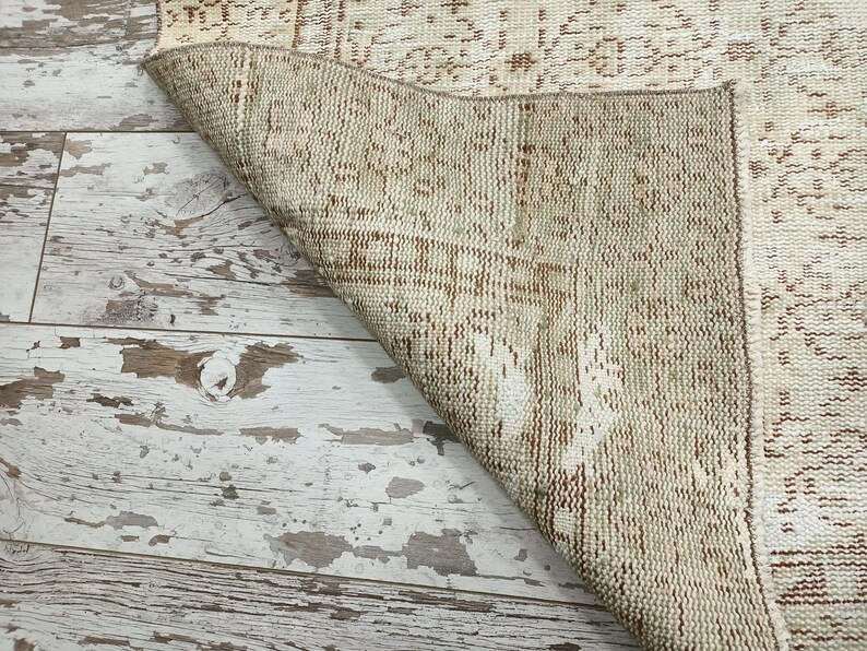 Puede incluir: Primer plano del reverso de una alfombra tejida beige y marr&oacute;n. La alfombra tiene un aspecto desgastado y est&aacute; doblada sobre un suelo de madera blanqueada.
