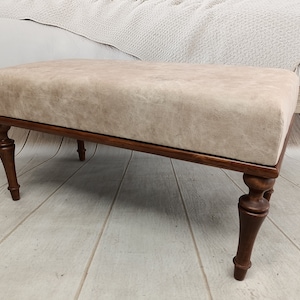May include: A beige upholstered bench with a wooden frame and turned legs. The bench is sitting on a wooden floor.