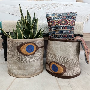 Puede incluir: Dos cestas de almacenamiento decorativas con un diseño de pluma de pavo real. Las cestas son beige con detalles marrones y ojos de pavo real azules y dorados. Una cesta está llena de una planta verde.