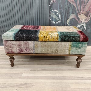 Puede incluir: Un otomano rectangular de patchwork colorido con patas de madera. El otomano está cubierto de diferentes telas en tonos de rojo, verde, azul, amarillo y marrón.