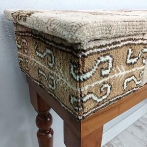 Op de afbeelding: Een houten kruk met een beige en bruin gepatroneerd stoffen bovenblad. De kruk heeft gedraaide houten poten.