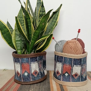 Könnte beinhalten: Zwei dekorative Stoffkörbe mit einem südwestlichen Muster. Einer enthält eine Bogenhanf-Pflanze, der andere Wollknäuel und Stricknadeln. Die Körbe haben ein Farbschema in Dunkelblau, Braun und Creme.