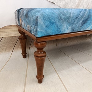 Puede incluir: Un otomano tapizado en azul y marr&oacute;n con patas de madera torneadas. El otomano tiene un patr&oacute;n abstracto azul y blanco en la parte superior.