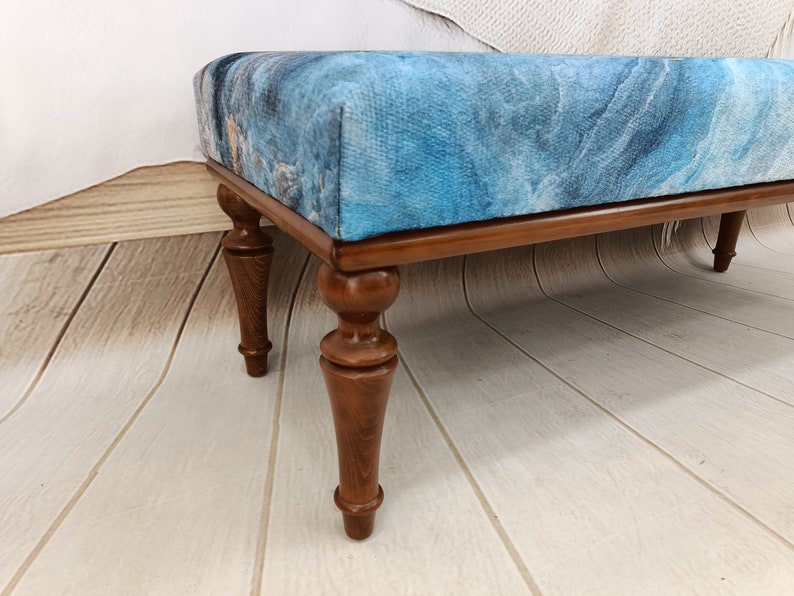 Puede incluir: Un otomano tapizado en azul y marr&oacute;n con patas de madera torneadas. El otomano tiene un patr&oacute;n abstracto azul y blanco en la parte superior.