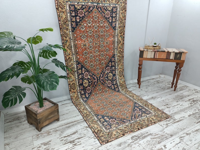 May include: A vintage rug with a floral pattern in shades of red, orange, blue, and cream. The rug has a fringed edge and is laid out on a whitewashed wooden floor.