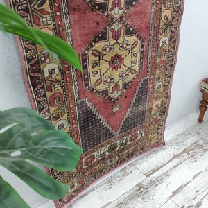 May include: A vintage Turkish runner rug with a red, cream, and black geometric pattern. The rug has a faded, distressed look and is perfect for adding a touch of bohemian style to your home.