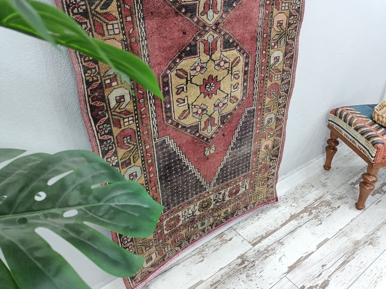 May include: A vintage Turkish runner rug with a red, cream, and black geometric pattern. The rug has a faded, distressed look and is perfect for adding a touch of bohemian style to your home.