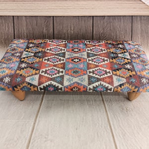 May include: A rectangular ottoman with a colorful geometric pattern in shades of red, orange, blue, and brown. The ottoman has wooden legs.