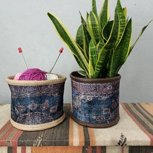 May include: Two fabric planters with a blue and pink patterned design. One planter holds a potted snake plant, and the other holds pink and white yarn with knitting needles.