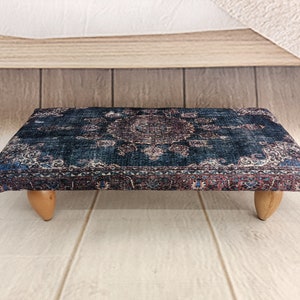 Puede incluir: Un otomano de color azul oscuro y rojo con estampado y patas de madera. El otomano está sobre un suelo de madera clara.