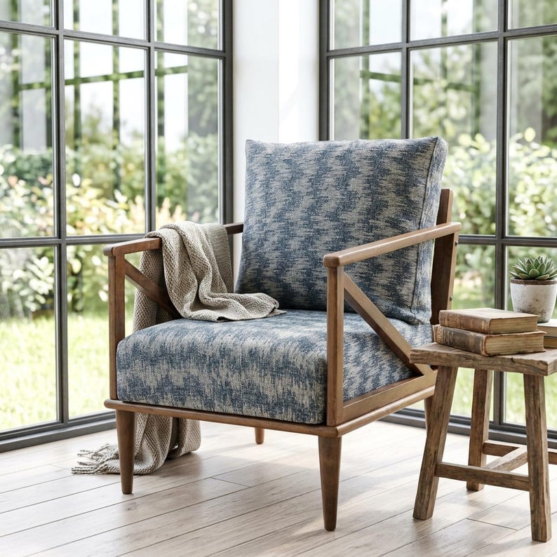 a cozy living room with a comfortable blue and white patterned armchair, a wooden side table, and a stack of books, all positioned near a large window that offers a view of a lush garden.