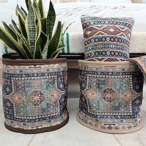 Puede incluir: Dos cestas de almacenamiento cilíndricas con cubiertas de tela estampada. Las cestas son beige y marrón con un patrón geométrico azul, verde y naranja. Una cesta tiene una planta dentro y la otra una almohada con un patrón similar.