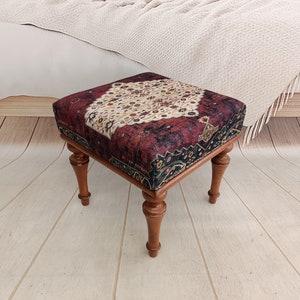 May include: A square ottoman with a red, black, and cream patterned fabric top and four wooden legs. The ottoman is sitting on a light wood floor.