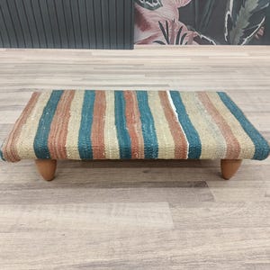 May include: A striped ottoman with wooden legs. The ottoman is covered in a fabric with stripes of beige, brown, teal, and white.