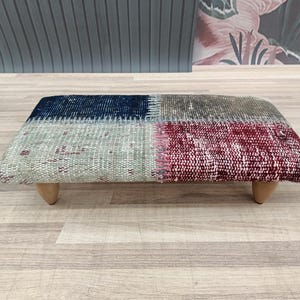 May include: A rectangular footstool with a patchwork design. The top features sections of blue, gray, and burgundy fabric. It rests on four short, light-colored wooden legs, offering a blend of textures and colors.