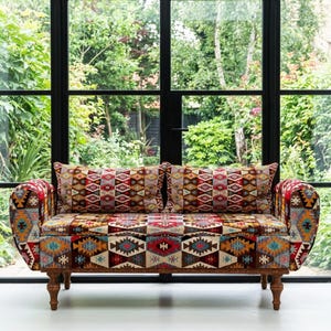 Può includere: Un divano a due posti con un design patchwork colorato. Il divano ha una struttura in legno e gambe tornite. Due cuscini abbinati con motivi geometrici poggiano sulla seduta. Il divano è posizionato davanti a una grande finestra.