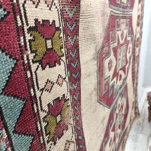 May include: A vintage Turkish rug with a cream background and a geometric pattern in shades of red, blue, and green. The rug has a frayed edge and is slightly worn.
