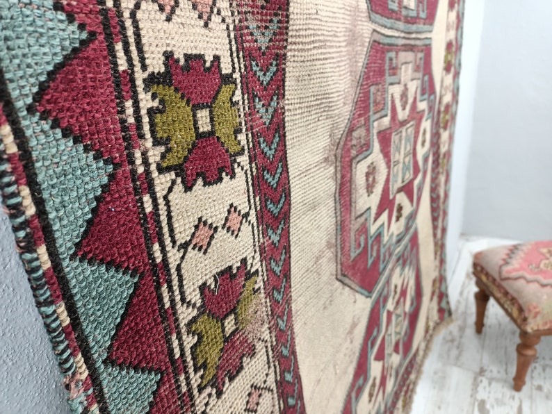 May include: A vintage Turkish rug with a cream background and a geometric pattern in shades of red, blue, and green. The rug has a frayed edge and is slightly worn.