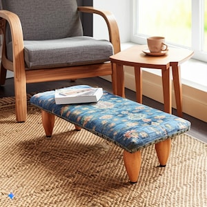 May include: A blue and brown patterned footstool with four wooden legs. The footstool is sitting on a woven rug in front of a wooden chair and a small wooden table.