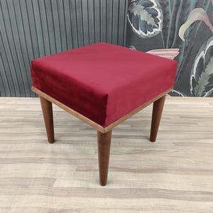 May include: A red velvet upholstered square ottoman with four wooden legs. The ottoman is sitting on a light wood floor.