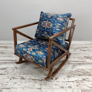 A wooden rocking chair with a blue and white patterned cushion, placed on a white wooden floor against a plain white wall.